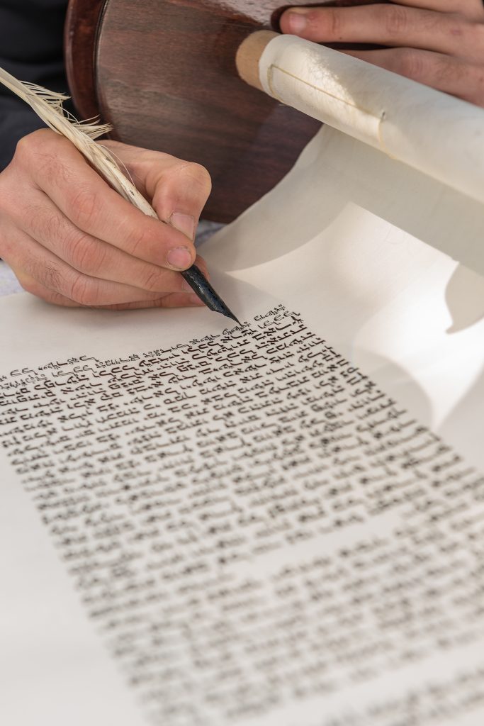 Persona escribiendo con una pluma de ave sobre un pergamino enrollado, con texto en caligrafía antigua.