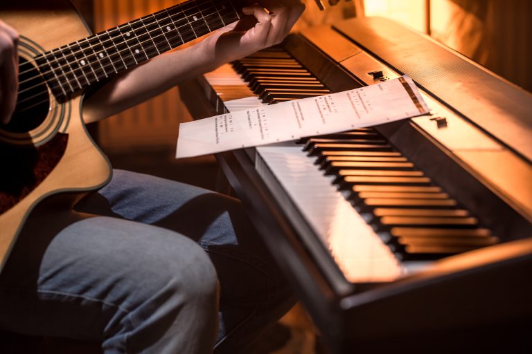 Persona tocando guitarra acústica junto a un teclado eléctrico con partituras apoyadas sobre las teclas.
