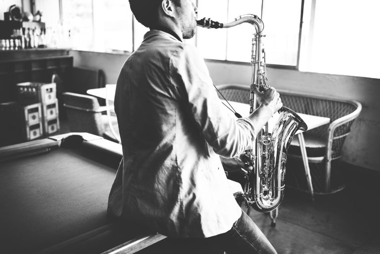 Músico tocando un saxofón en blanco y negro, sentado sobre una mesa de billar en un ambiente interior con luz natural.
